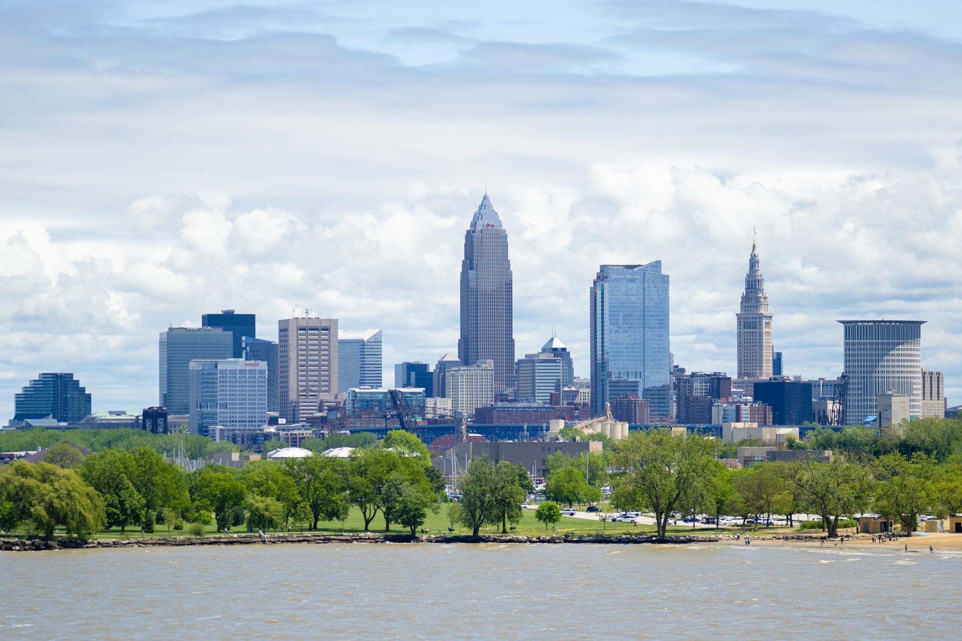 Cleveland skyline across the river with green trees in the foreground under a cloudy sky