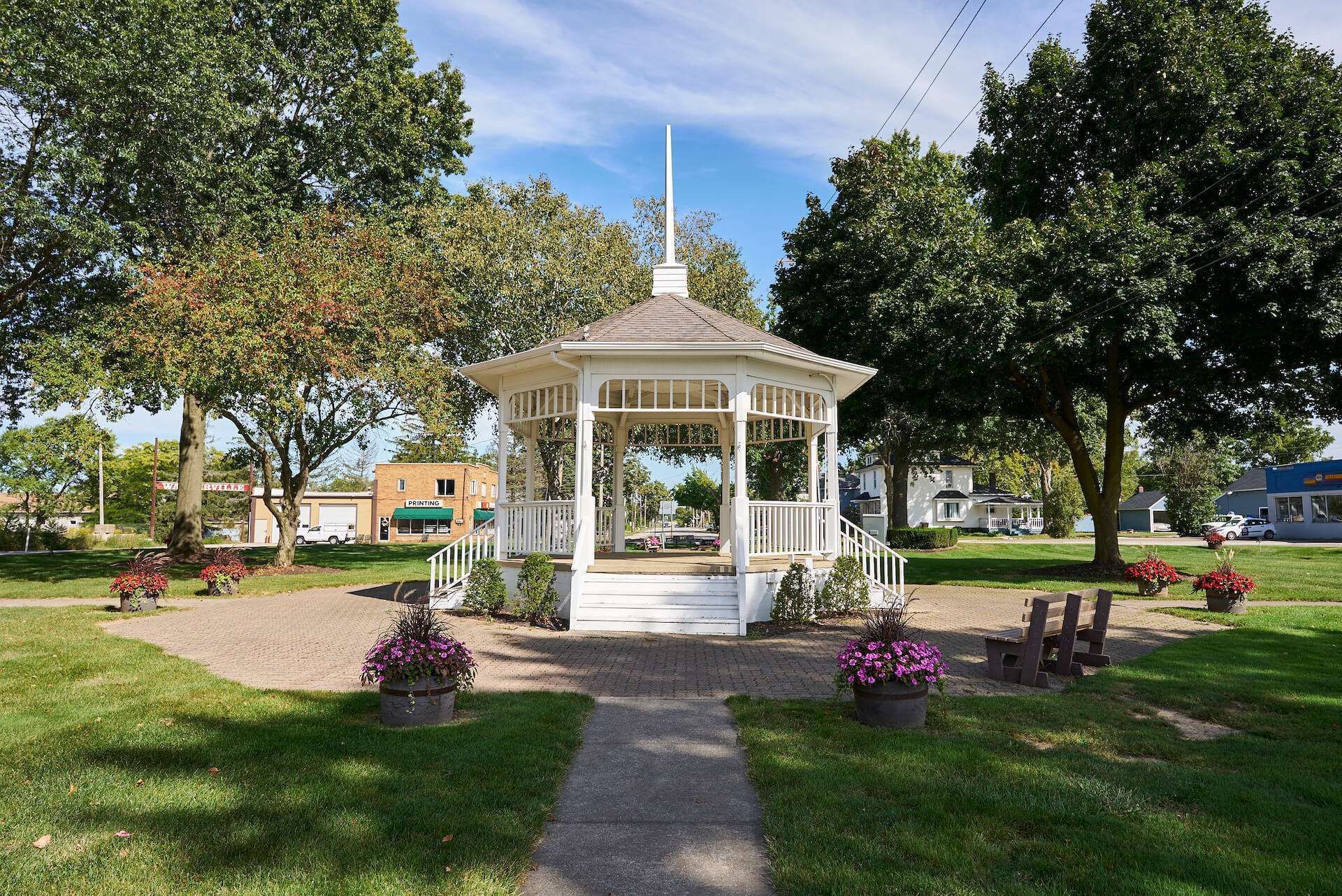 White gazebo in a sunny park with green trees, grass, and flower pots along a paved path.