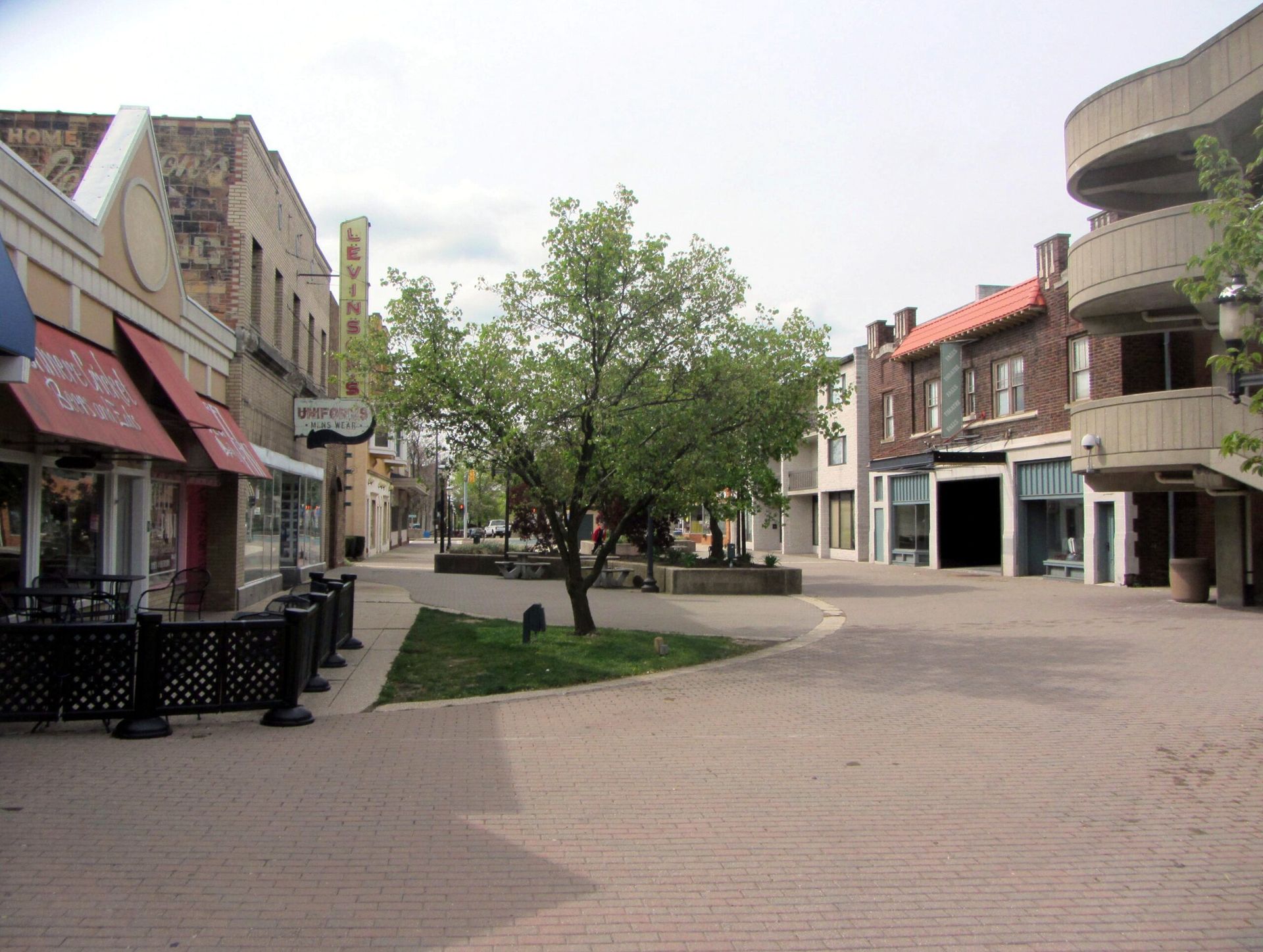 Quiet brick-paved street with storefronts, a central tree, and a few parked cars in a small town square