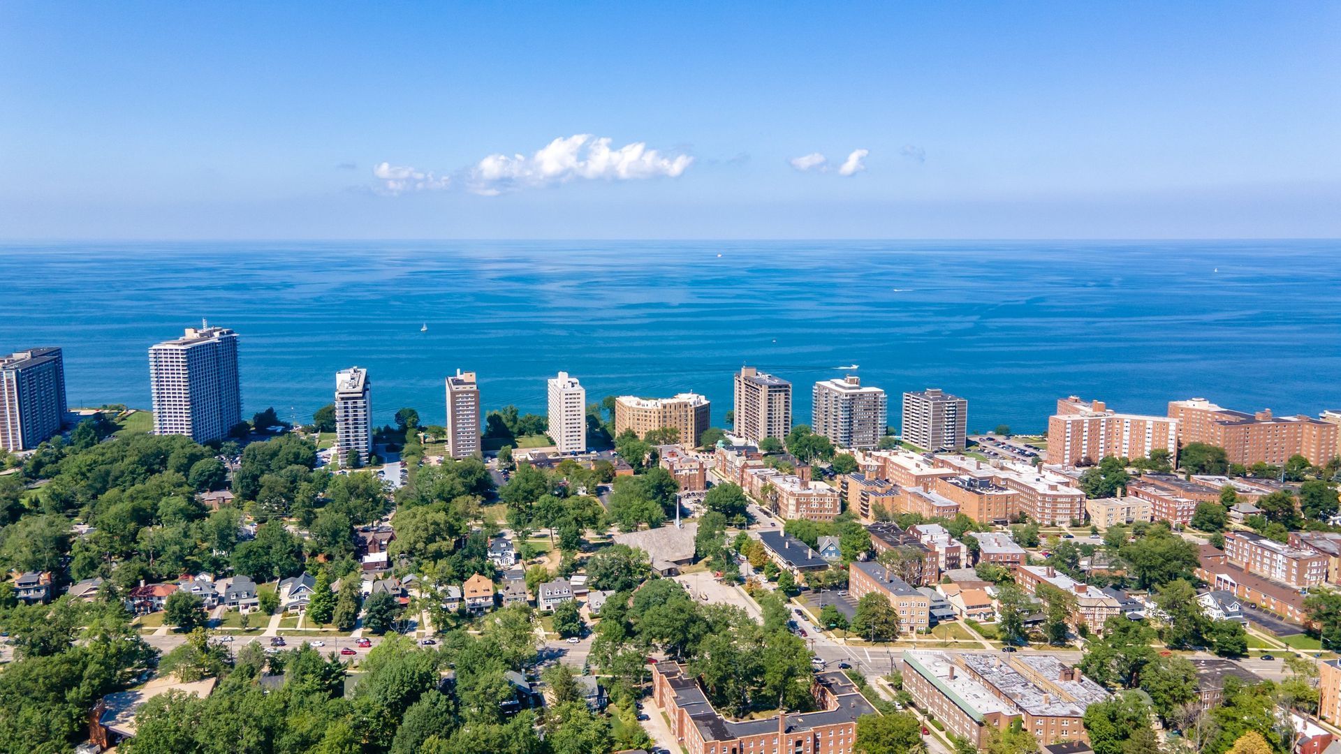 Coastal city skyline with apartment buildings, trees, and a calm blue ocean under a clear sky