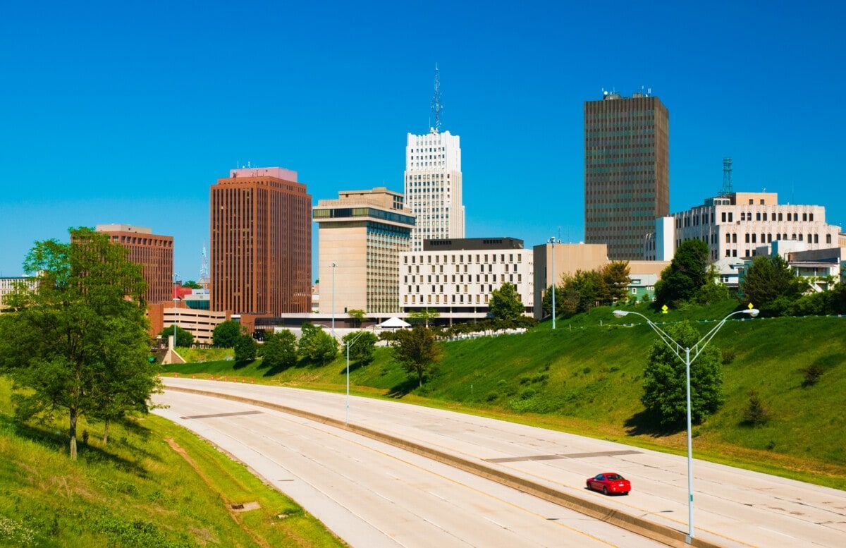 City skyline with tall buildings beside a highway, green hills, and a red car under a blue sky