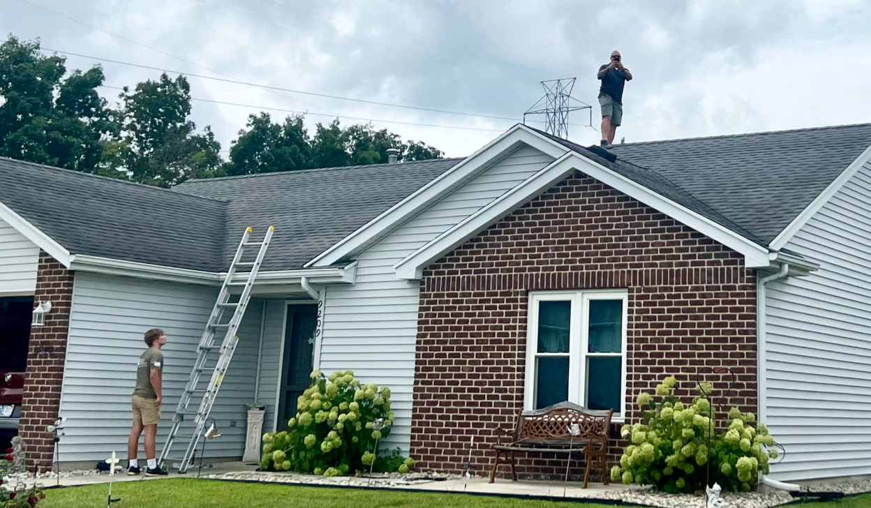 Two people are working on the roof and siding of a suburban house with a brick and gray horizontal siding exterior.