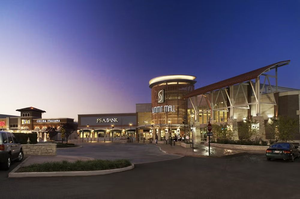 Dusk view of a modern shopping center with illuminated storefronts, glass facade, and parked cars.