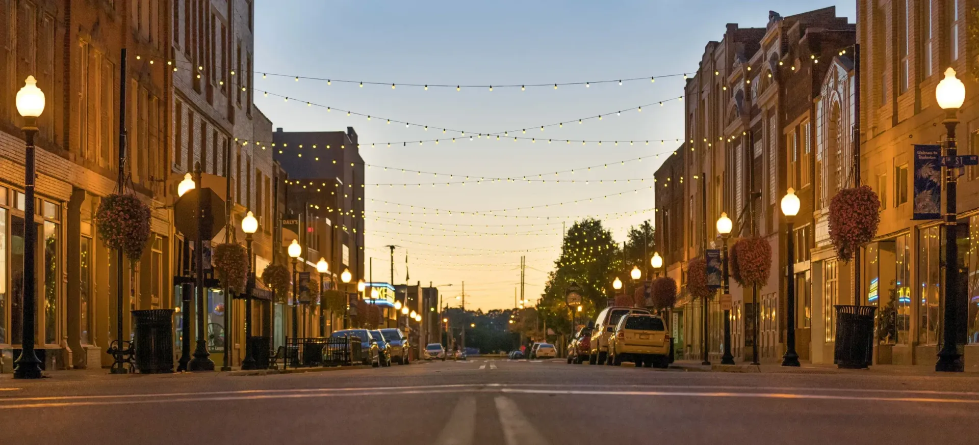Sunset view of a quiet city street lined with lit historic buildings and parked cars
