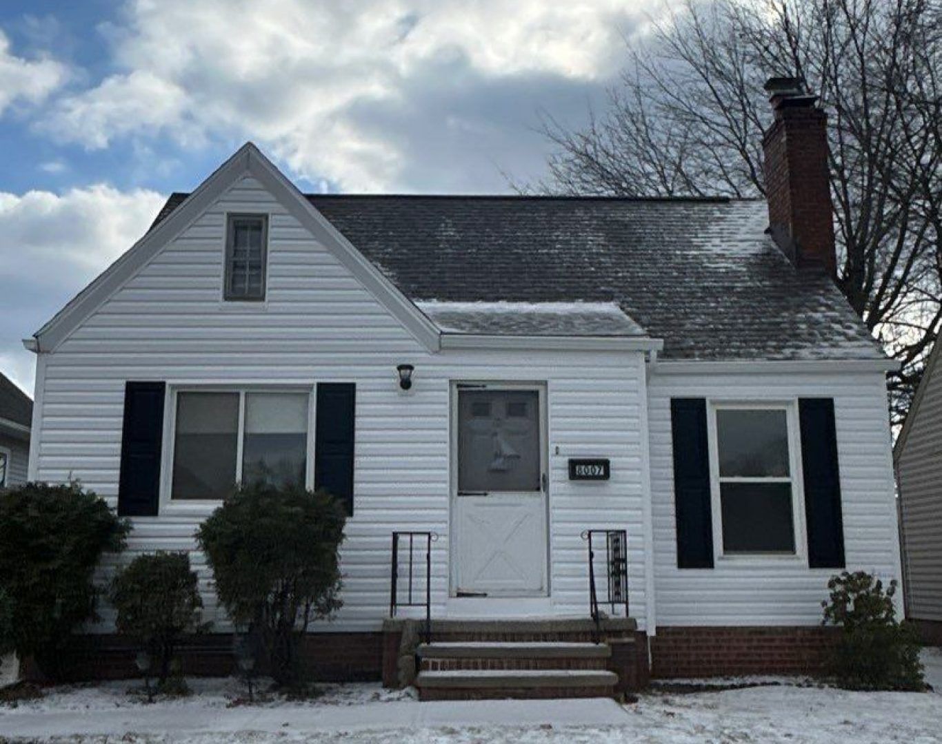 Small white house with black shutters, front steps, and a brick chimney on a cloudy winter day