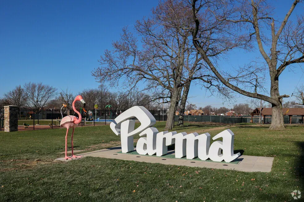 Pink flamingo beside large white “Parma” sign in a grassy park under a blue sky.