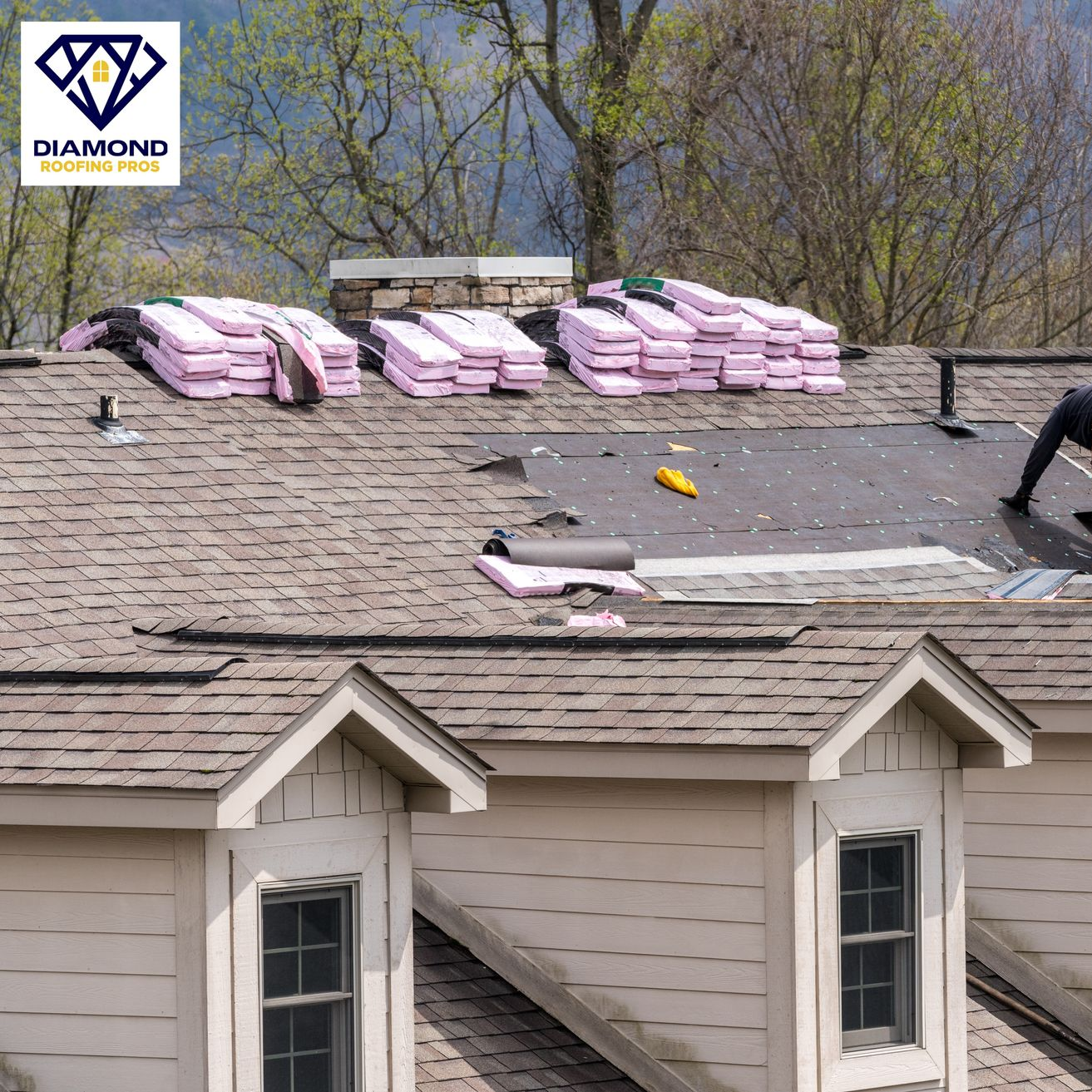 Worker repairing a shingled roof beside stacked pink insulation on a house