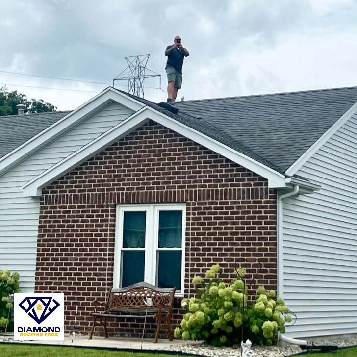 Worker standing on a shingled roof above a brick house, inspecting it.