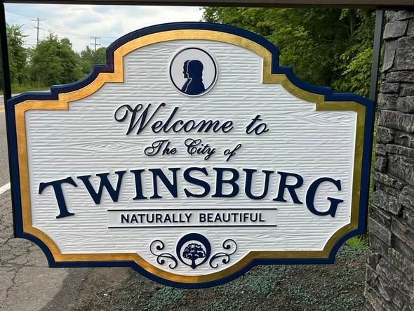 Welcome to Twinsburg city sign, white with blue and gold trim, outdoors against trees and stone wall