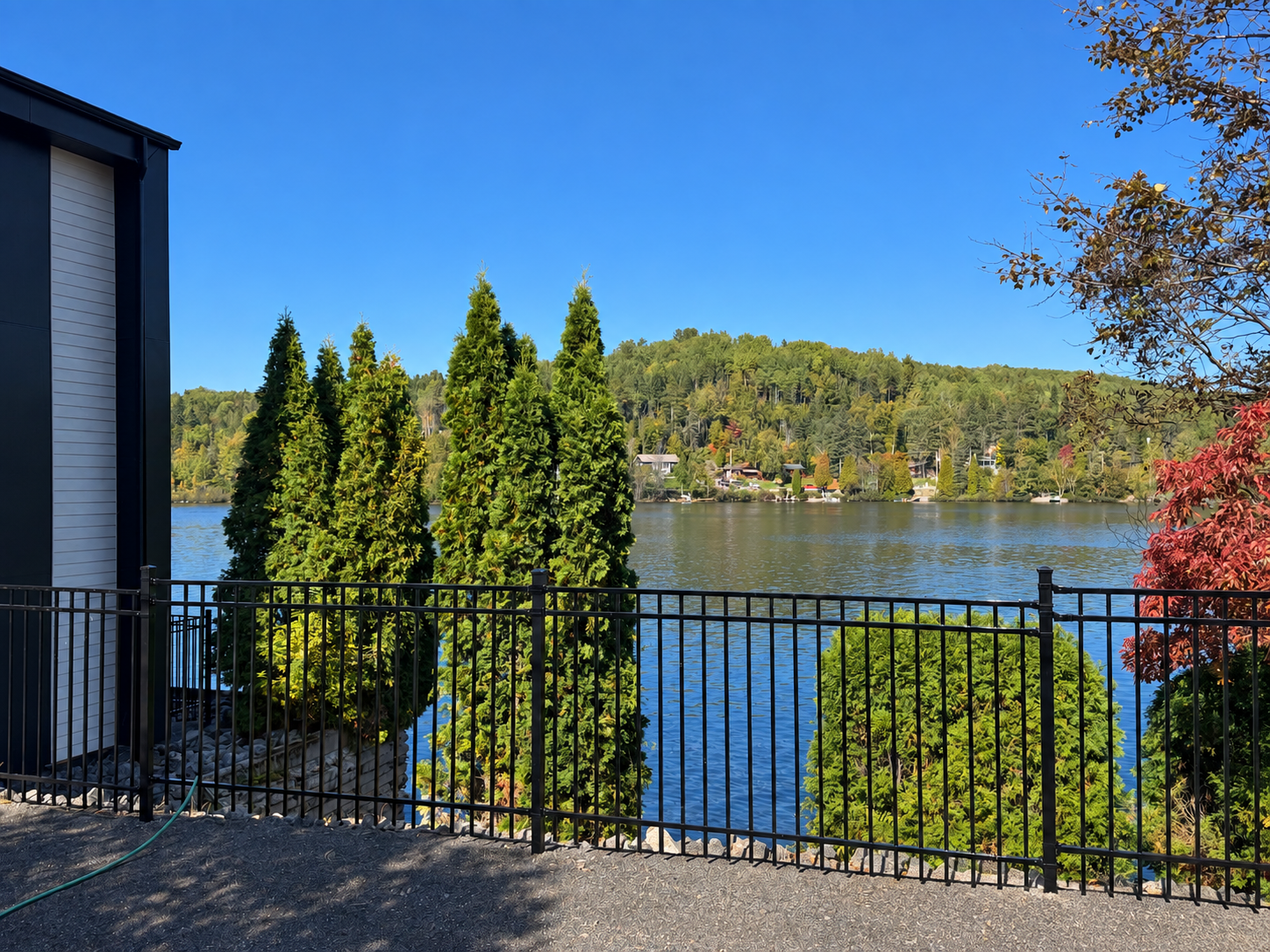 Vue sur le lac depuis un balcon avec une rambarde noire, des conifères et un ciel bleu.