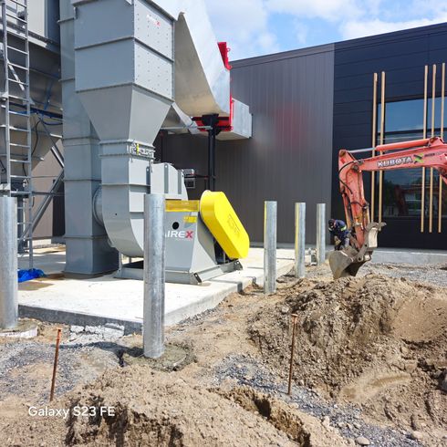 Chantier de construction avec une excavatrice, de grandes gaines argentées et une machine jaune à côté d'un bâtiment en construction