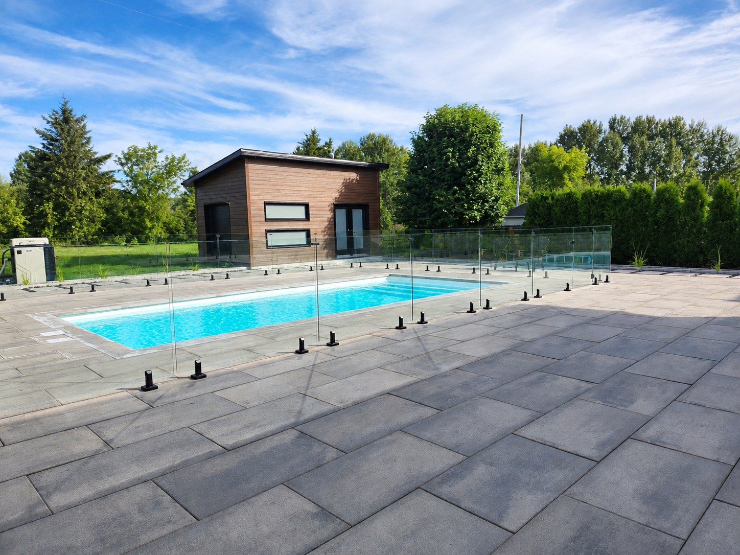 Piscine extérieure à côté d’un petit bâtiment en briques et d’une terrasse pavée, entourée d’arbres et de pelouse.