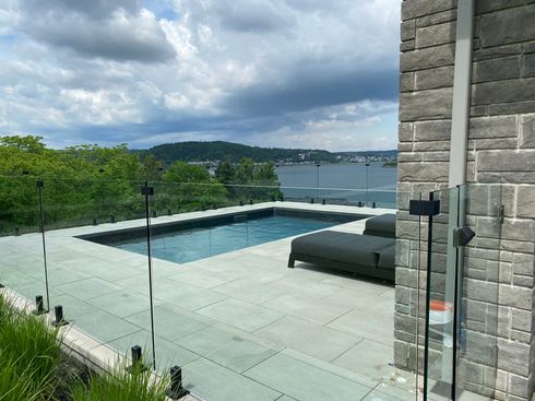 Patio moderne avec piscine en verre, mur en pierre, chaise longue et vue sur le lac nuageux