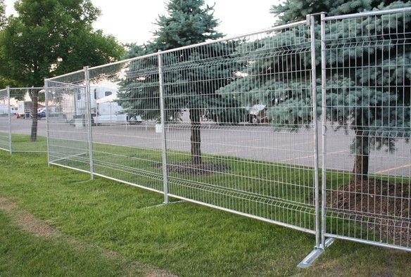 Des panneaux de clôture métalliques temporaires longent une route herbeuse, près des arbres et des maisons.