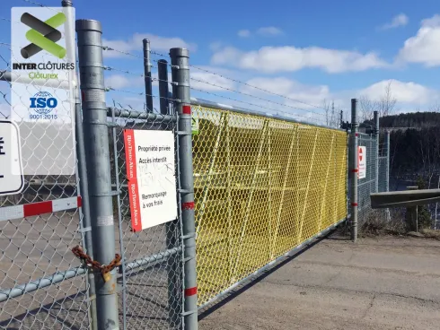 Clôture en grillage autour d'un site industriel, avec panneaux d'avertissement et filet de sécurité jaune.