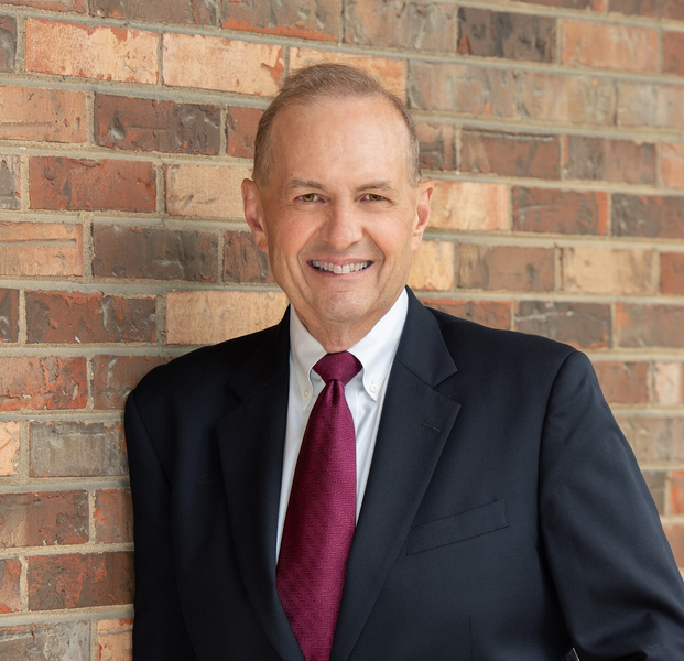 Smiling man in a dark suit and burgundy tie standing against a brick wall