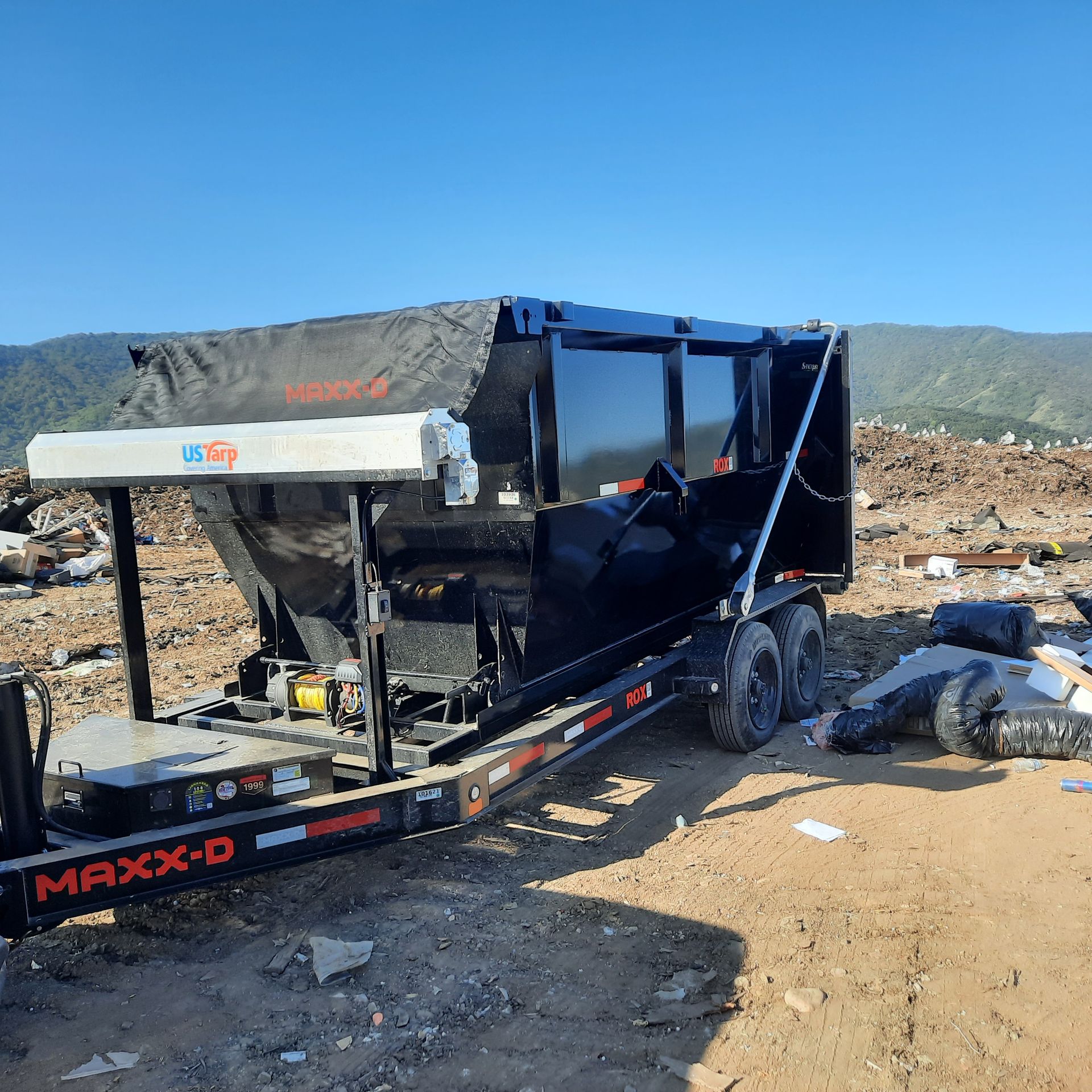 Black dump trailer on a dirt lot, mountains in the background, bright blue sky.