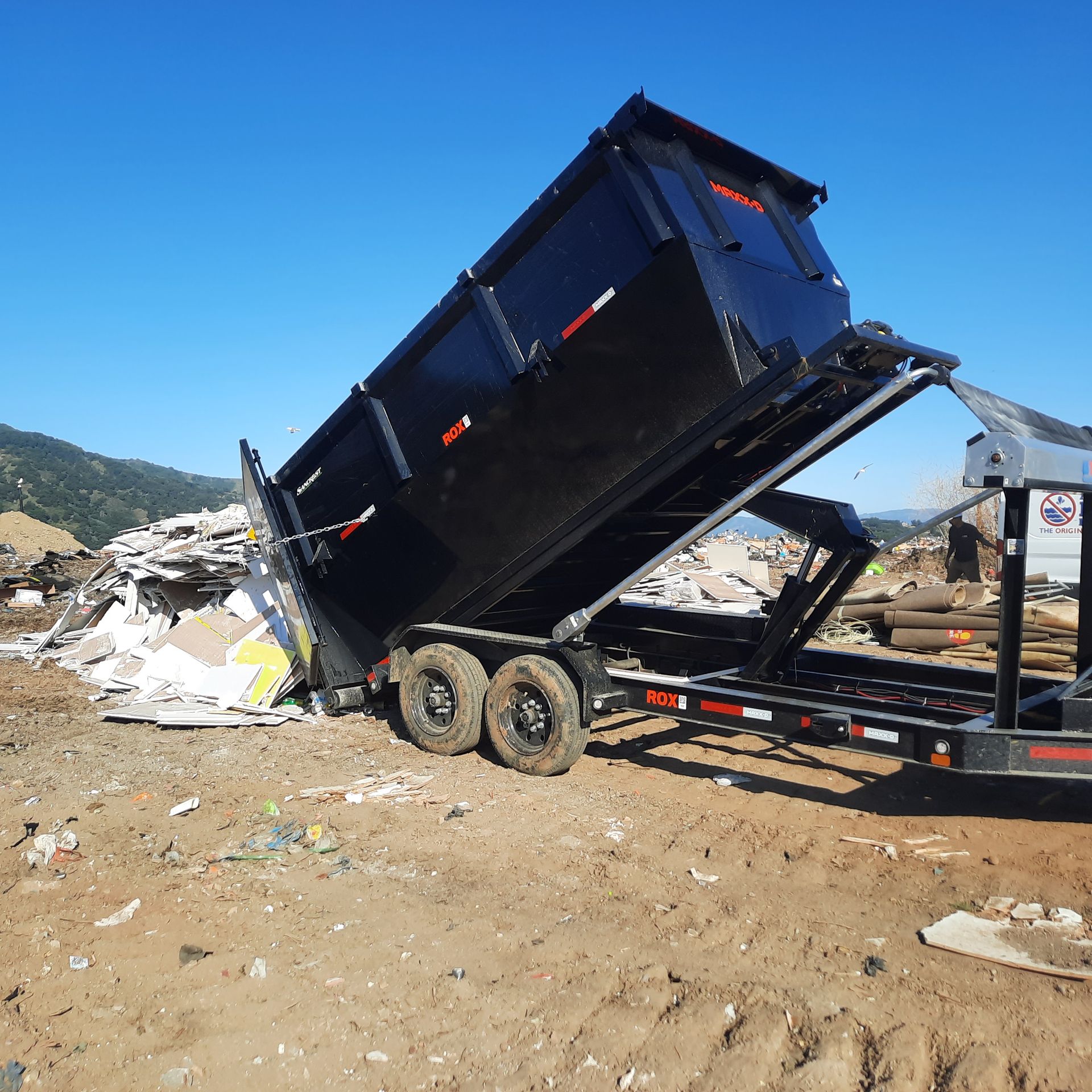 Black dumpster trailer, tilted, dumping debris onto a dirt ground, under a blue sky.
