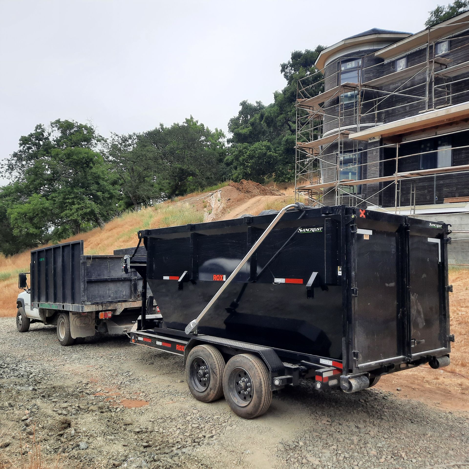 Black dump trailer attached to a truck on a construction site. A house with scaffolding is in the background.