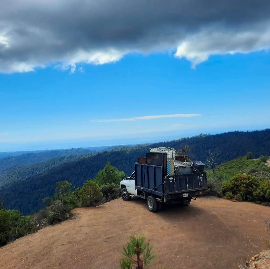 Truck loaded with debris parked on a ridge overlooking a forested valley under a partly cloudy sky.