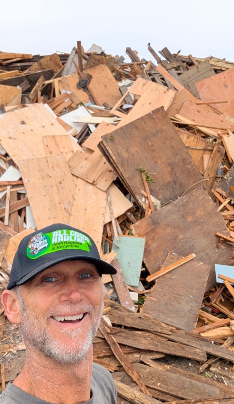 Man smiling in front of a large pile of construction debris; wearing a cap.