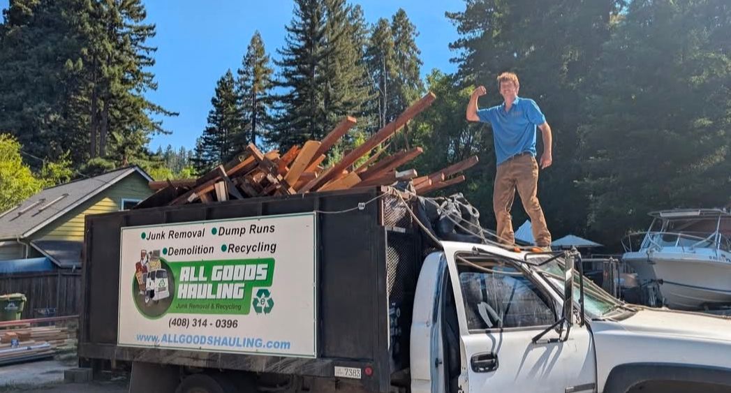 Man standing on a truck bed filled with wood debris; sunny outdoor setting.