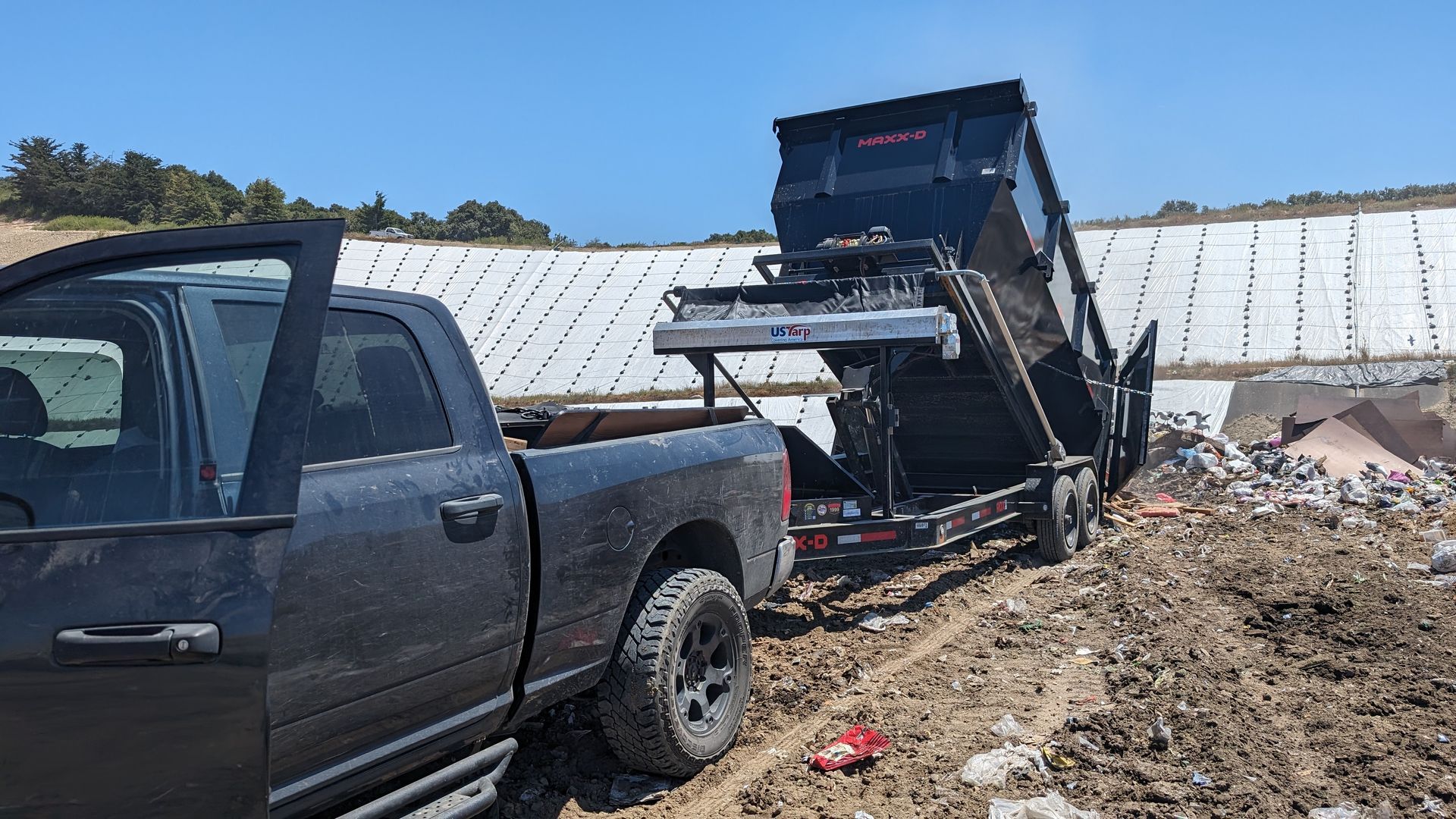 Black pickup truck towing a dump trailer at a landfill, dumping debris on a sunny day.