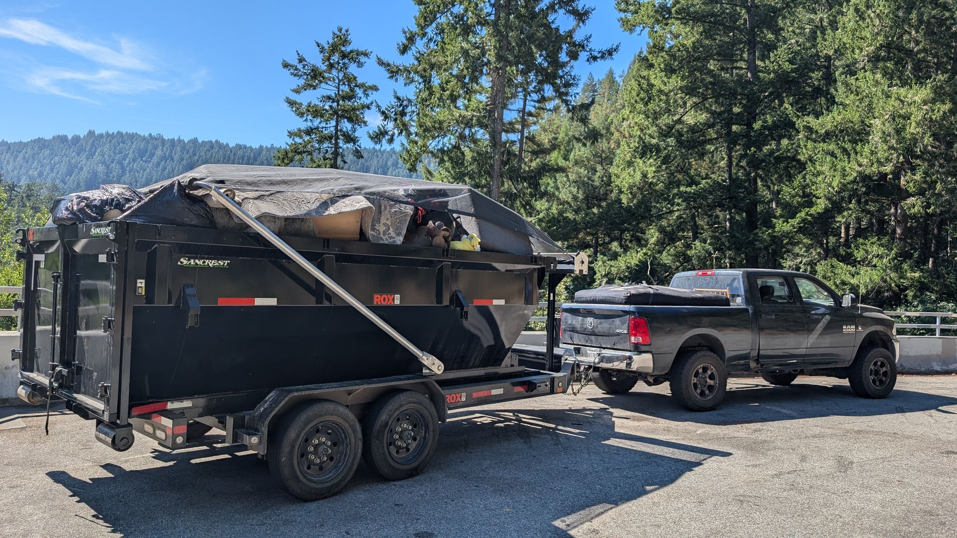 Black truck towing a black dumpster trailer with debris, parked on gravel with forest backdrop.