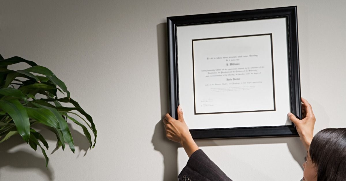 A woman in a dark blazer is hanging a certificate in a black frame on a gray wall. A tall, green pla