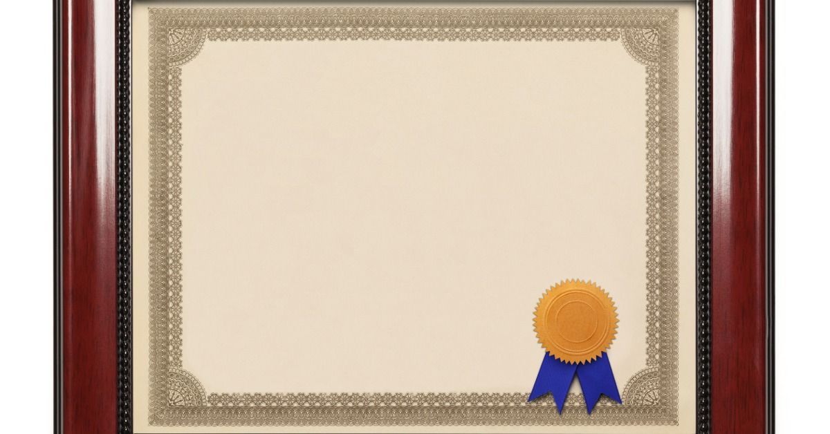 A blank certificate with a thick, ornate green border and a gold seal with blue ribbons is inside of a dark wood frame.