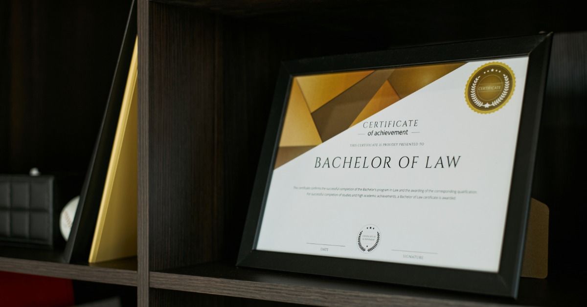 A bachelor of law certificate in a black holder is leaning on a dark wooden shelf beside gold folders and books.
