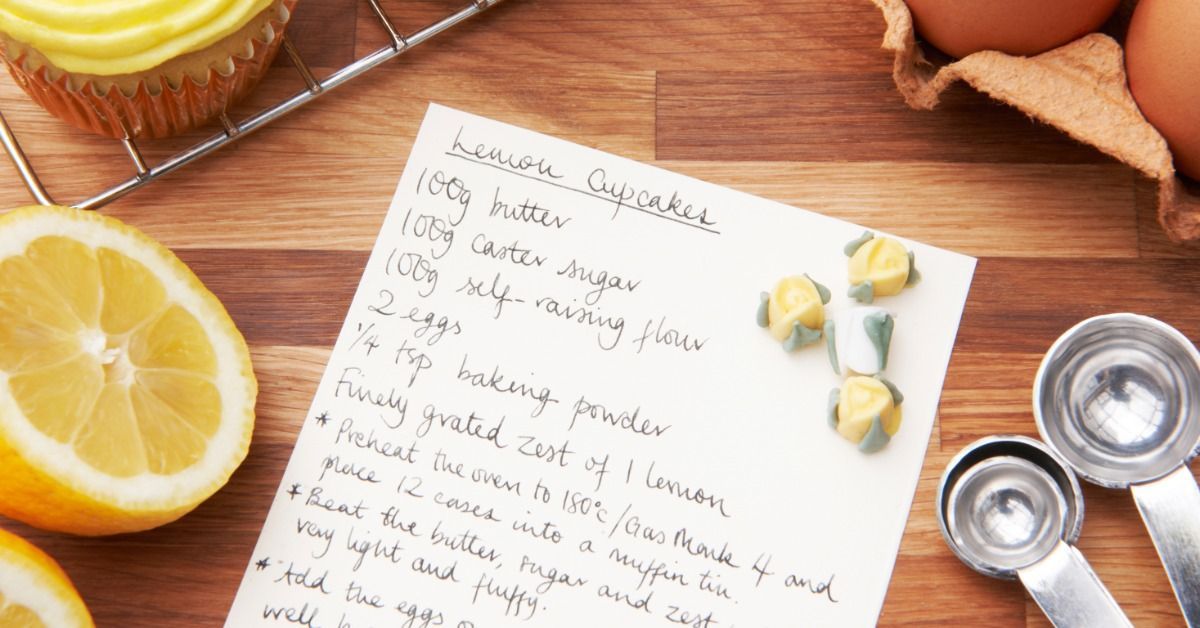 A handwritten lemon cupcake recipe lies on a wooden table beside yellow frosted cupcakes, lemons, eg