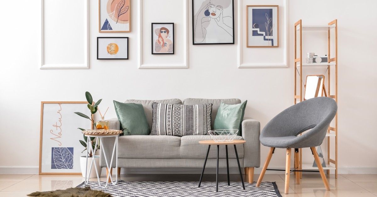 A combination of white, black, and tan picture frames are arranged on a white wall above a small gray couch.