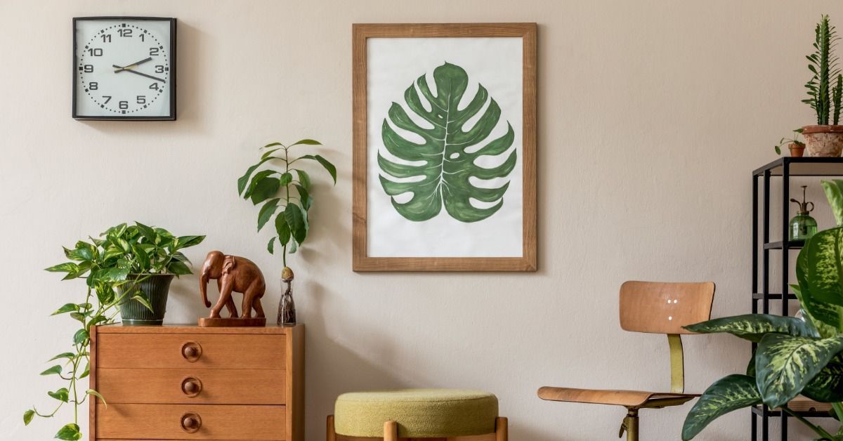 A room with a small storage table, cushioned chair, and potted plants. A framed poster of a monstera leaf is on the wall.