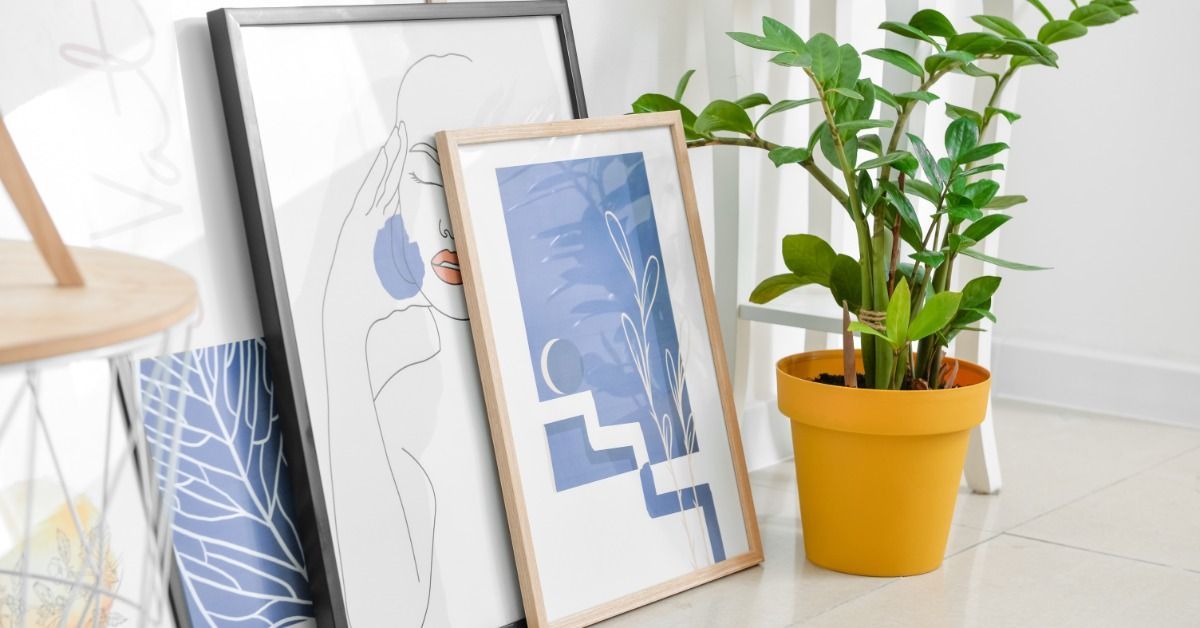 Several framed pieces of art are resting against a wall. A tall green houseplant in a yellow pot sits beside the art.