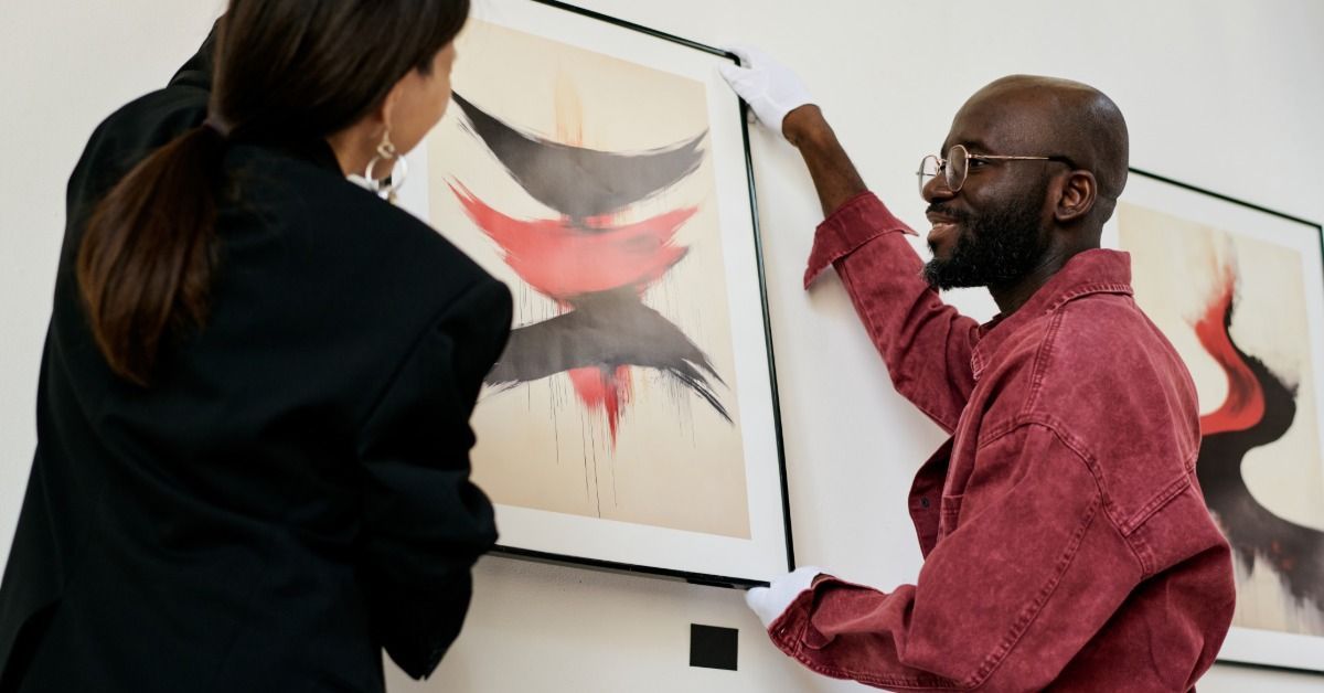 Two people wearing white gloves are hanging up a framed piece of art in a gallery. The art is painted red and black.