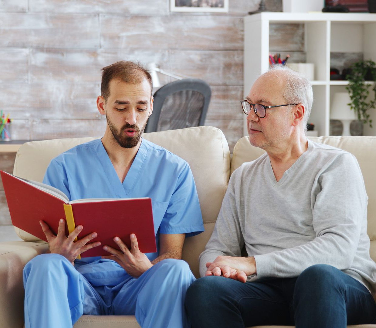 Caregiver in blue scrubs reads to older person on a couch. Crutch beside them. Caregiver in blue scrubs reads to older person on a couch. Crutch beside them.