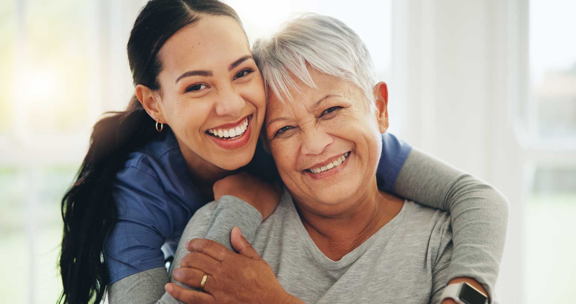 A smiling nurse, in a blue uniform, hugs a senior patient, showcasing home health care service.