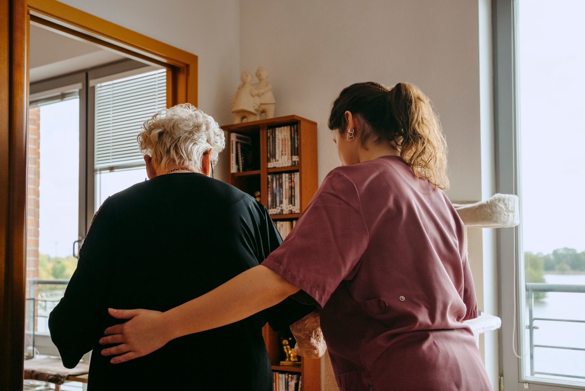 Caregiver helping elderly person in room with bookshelf and window.