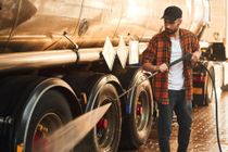 Man power washing a truck's tires with a hose, wearing a plaid shirt and a black cap, outdoors.