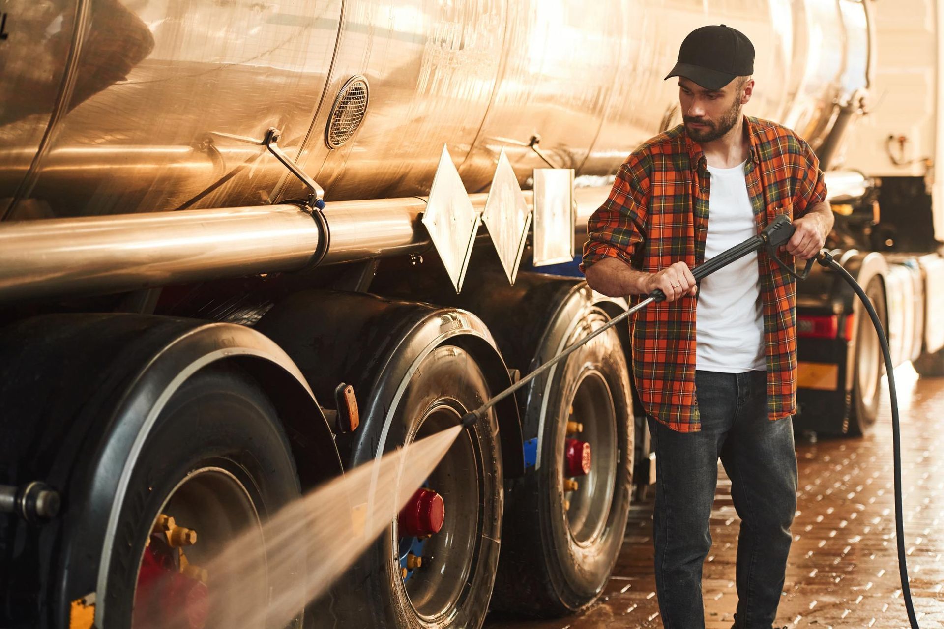 Man washing truck tires with a pressure washer in a garage, wearing a cap and plaid shirt.