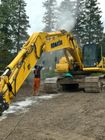 A worker in orange coveralls washes a large yellow Komatsu excavator with a pressure washer; trees in background.
