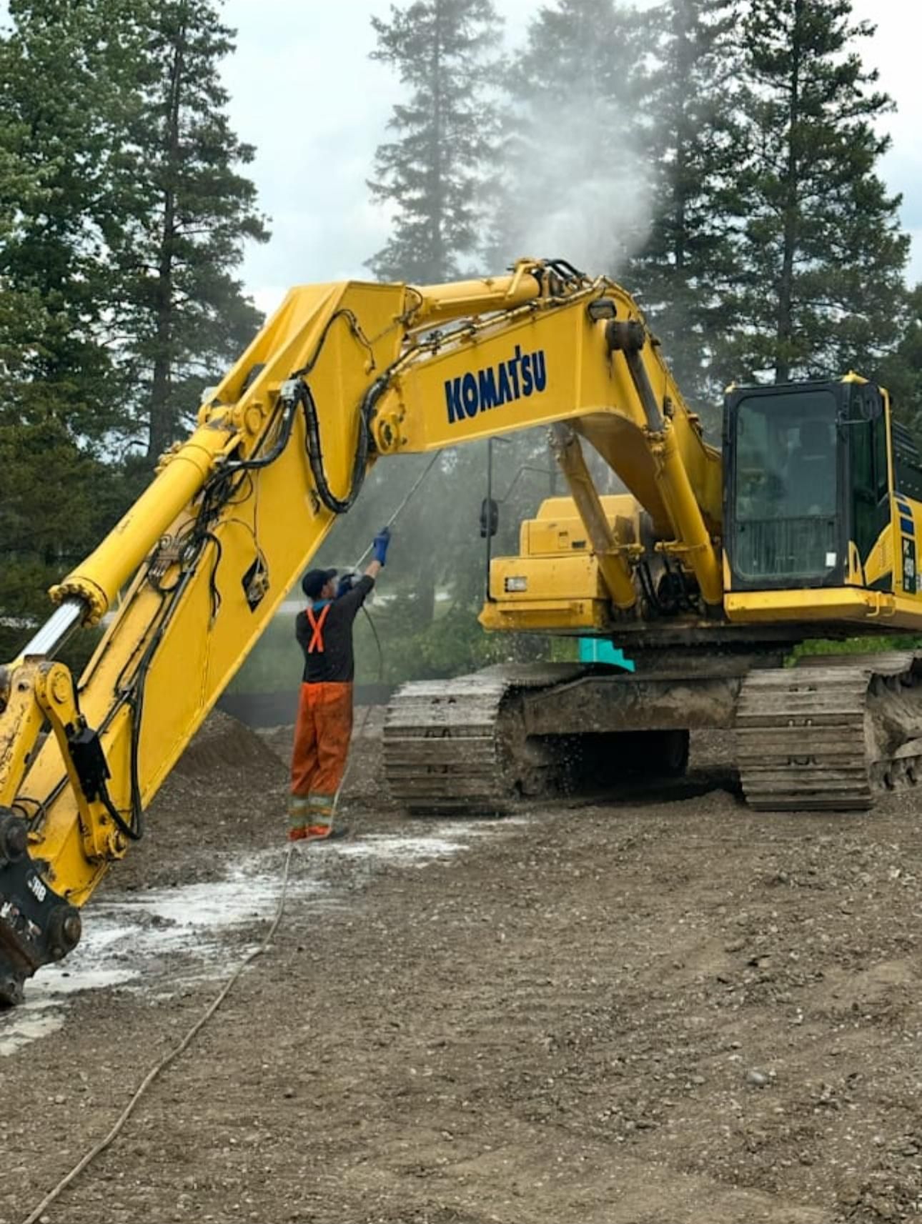Yellow Komatsu excavator being washed by a person in orange coveralls in a gravel area.