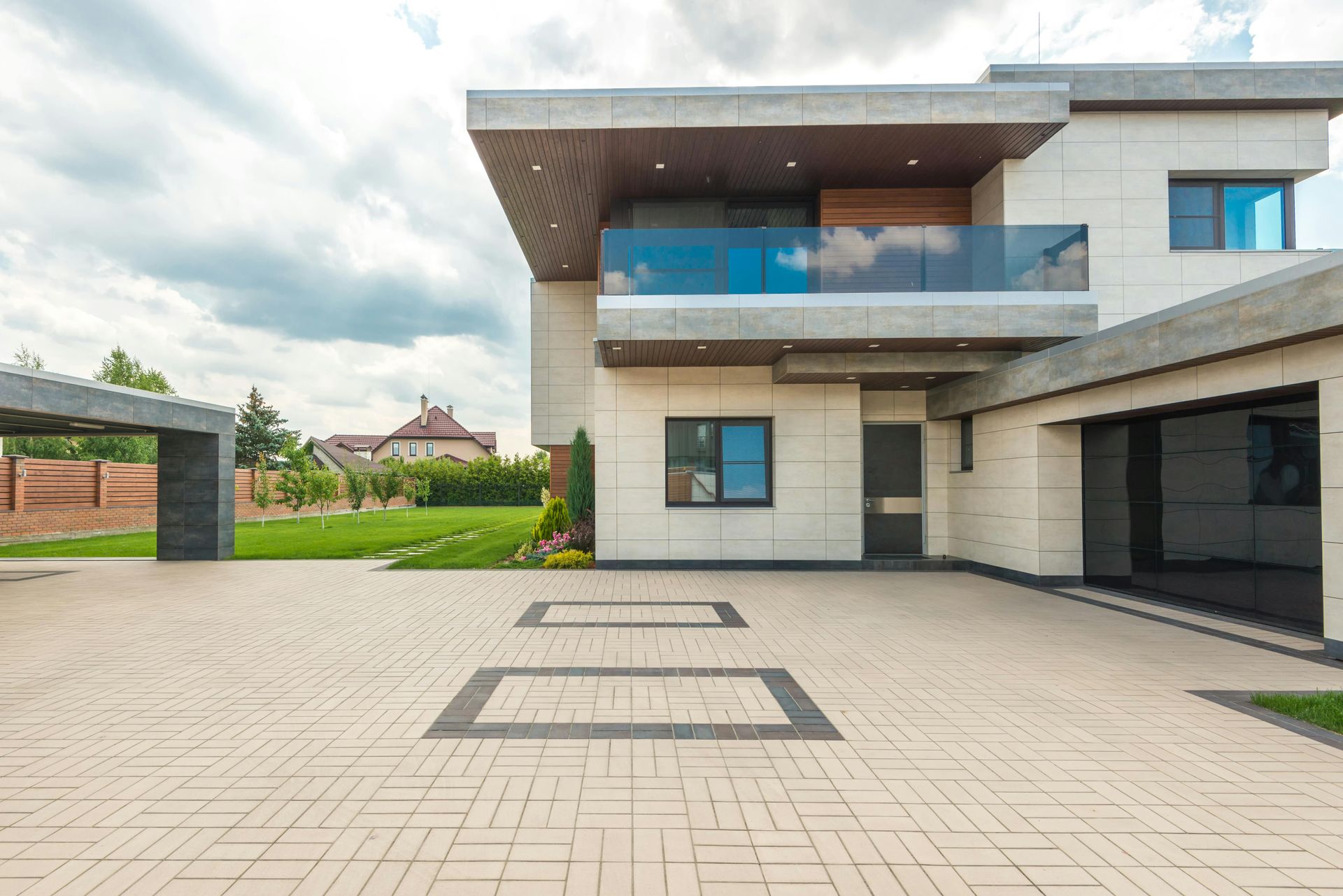 Modern two-story house with a light-colored exterior, glass balcony, and brick driveway under a cloudy sky.