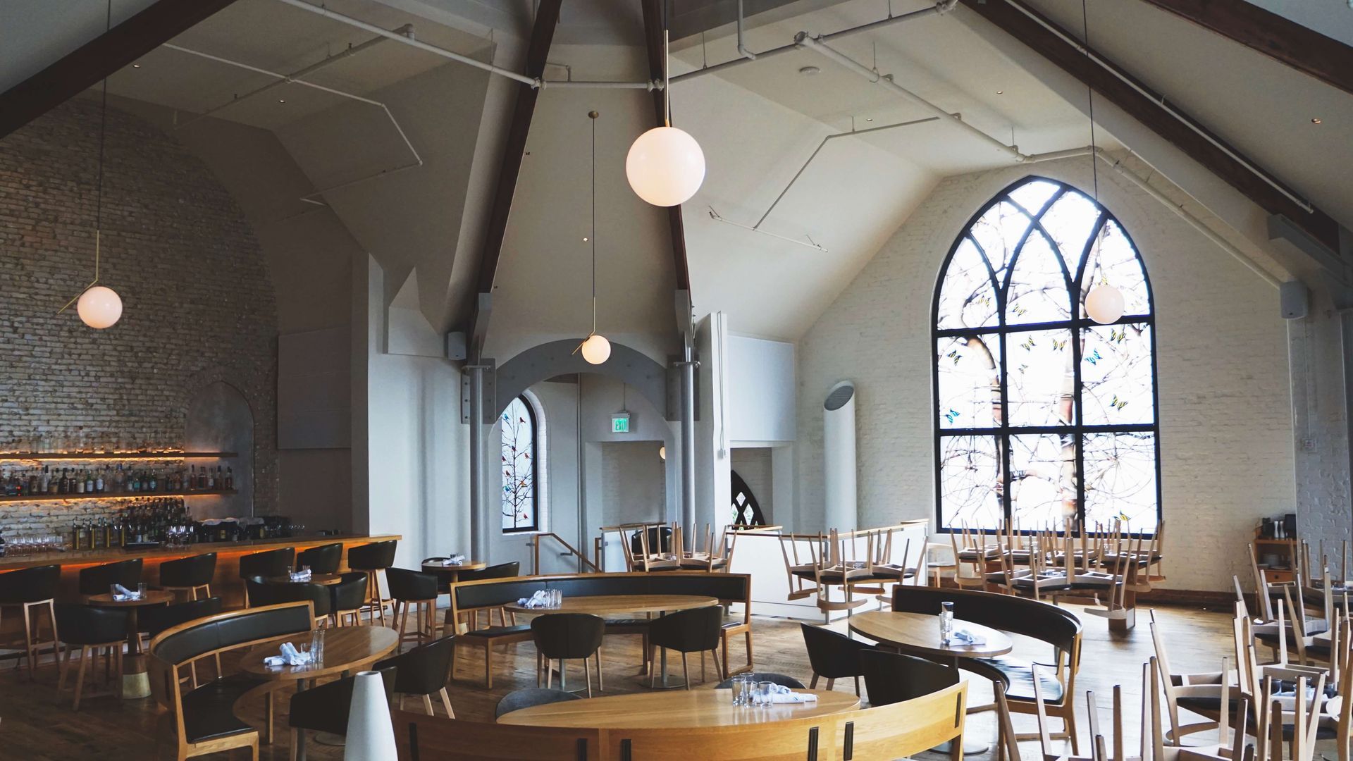 Restaurant interior in a former church with arched windows, round tables, and globe lights. Bar on the left.