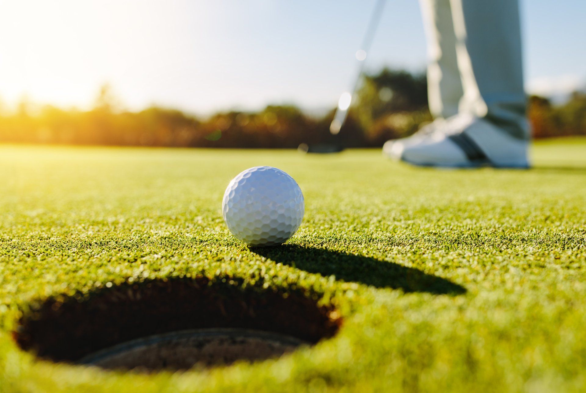 Golf ball near the hole on a green, with a golfer putting in the background under the sunlight.