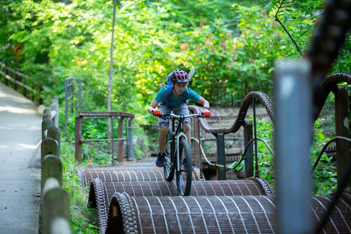 A person rides a mountain bike over a bridge in a wooded area, wearing a helmet and looking forward.