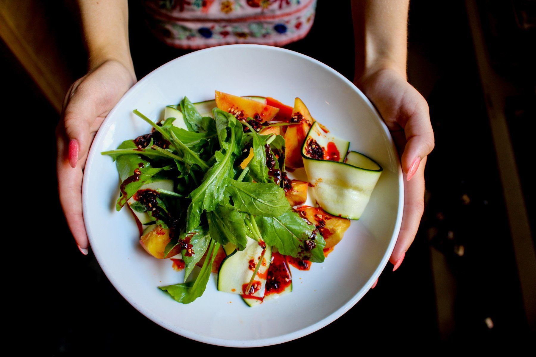 Hands holding a white bowl filled with a salad of green leaves, sliced vegetables, and red dressing.