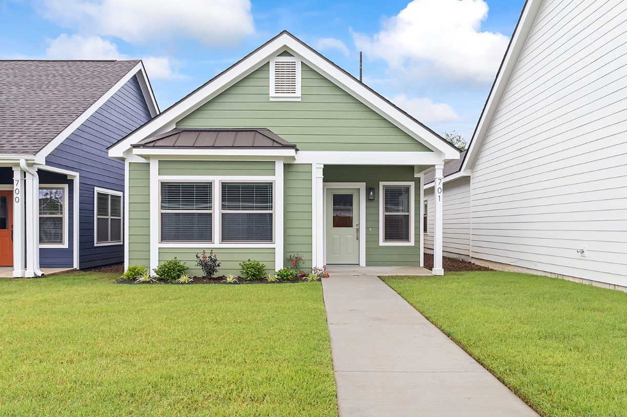 Green single-story house with white trim, gray roof, and walkway. Set between houses painted blue and white.