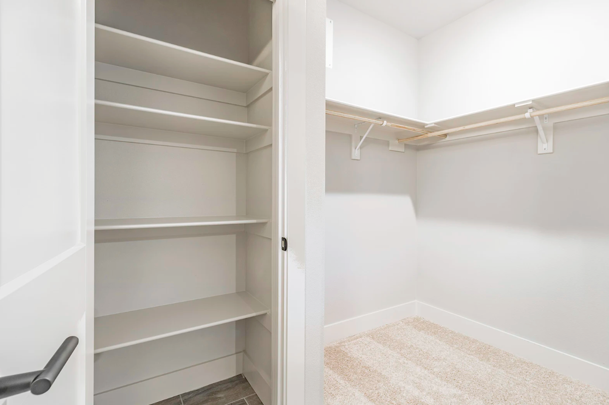 A white walk-in closet with shelves on one side and rods on the other. Carpeted floor.